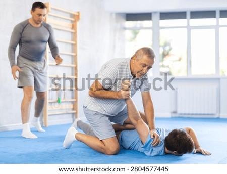 Determined elderly man attending self-defense classes at training center, applying arm lock technique to male opponent in mock bout
