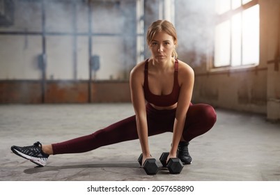 Determined Female Athlete With Dumbbells Looking At Camera While Doing Side Squat During Training In Gymnasium