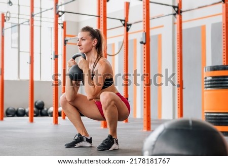 Determined female athlete looking away and doing goblet squat with heavy kettlebell during intense training in spacious light gym