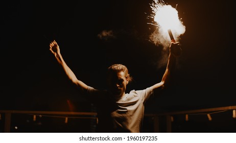 Determined Guy Standing With Fire On Roof. Fanatical Man Waving Torch Outdoors. Aggressive Football Fan Signaling With Flare Outside. Male Protester Burning Light In Urban Background. 