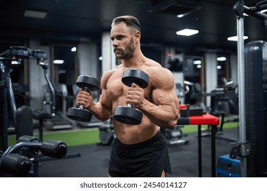 Determined handsome male weightlifter working out in gym. Portrait of bearded man with hairstyle, lifting dumbbells and looking aside, against blurred background. Bodybuilding, sport concept. - Powered by Shutterstock