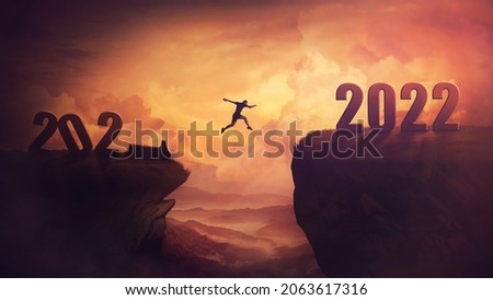 Determined man jump over a chasm obstacle to reach the new 2022 peak and let 2021 behind. Conceptual and surreal sunset scene, new year motivational background. Leader overcoming hurdles reach highs