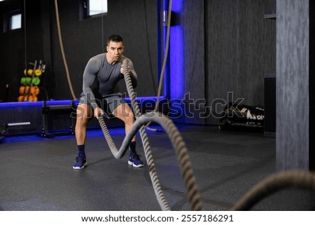 A determined man performs a high-intensity battle rope exercise in a modern gym, displaying strength and endurance amidst a motivating athletic environment.