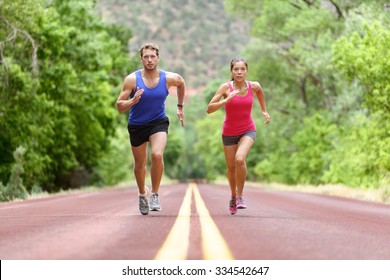 Determined Man And Woman Running On Road Against Trees. Runners Sprinting In Full Length Of Sporty Male And Female Are In Sports Clothing. Athletic Runner Fitness Sport Couple Are Exercising Outside.