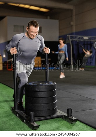 Determined middle-aged man pushing heavily loaded sled across gym floor, engaging in high-intensity functional workout..