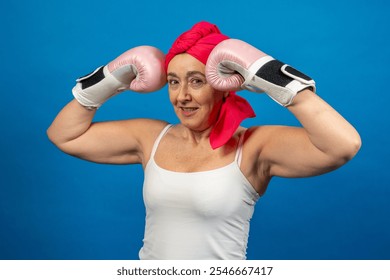 Determined senior woman wearing pink boxing gloves and red headscarf flexing her biceps on blue background - Powered by Shutterstock