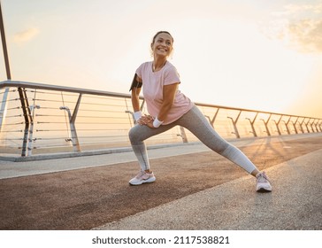 Determined sportswoman with healthy body doing side lunges, stretching her legs muscles while working out outdoor on a city bridge at dawn. Sport, fitness, body care and active lifestyle concept - Shutterstock ID 2117538821