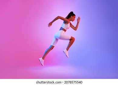 Determined Sporty Woman Running In Mid-Air Exercising During Cardio Workout Over Pink And Blue Neon Studio Background, Wearing White Fitwear. Fitness And Sport Concept. Side View, Full Length