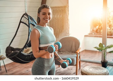 Determined woman losing weight at home and exercising with dumbbells. Sport and recreation concept. Beautiful woman in sportswear with blue dumbbells in her hands.