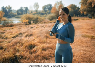 A determined young adult female athlete adjusting her earphones, holding a mobile phone and preparing for her routine outdoor workout. - Powered by Shutterstock