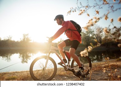 Determined Young Bearded Man In Long Sleeve Jersey Riding A Mountain Bike By The Lake Or River. Sunset Over Water In Background. Side View