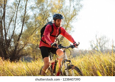 Determined Young Bearded Man In Red Long Sleeve Cycling Jersey Riding Mountain Bike Along A Path Through Tall Grass. Low Angle Front Side View.