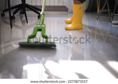 determined young boy in yellow boots is working hard to restore his home after a flood. Armed with a mop he bravely battles against the mud and water to make his living space habitable again.