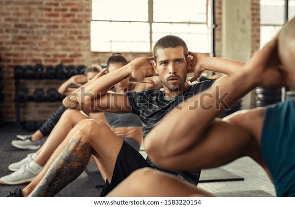 Determined young man\
doing exercise for abdominal. Athletic class doing abs in a row.\
Fitness man in gym exercising while sitting on mat in fitness\
center during cardio\
course.