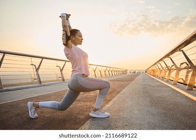 Determined young sportswoman exercising outdoor at dawn. Female athlete in sportswear stretching her arms up, performing squats and lunges, working out on city bridge in the early morning at sunrise. - Shutterstock ID 2116689122