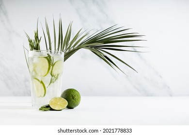 Detox Cold Lime Lemonade With Ice Cubes, Lime Slices, Rosemary Twig, Ingredients, Green Palm Leaf  In Transparent Glass On White Wood Table In Sunlight, Summer Cocktail.