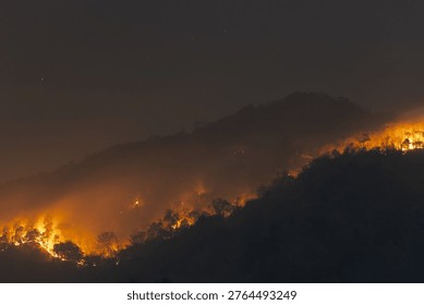 Devastating massive wildfire burning on mountain ridge at night emitting heavy smoke pollution into dark sky causing terrifying environmental disaster near Chiang Mai Thailand