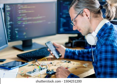 Developer is connecting breadboard to microcontroller. Man is holding smartphone with program code software for controlling electronic device. Chips, resistors, diodes on desktop of hardware engineer.