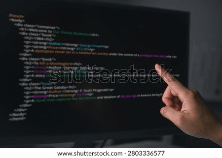 Developer hand pointing directly at a computer screen displaying colorful HTML programming code, representing software development, website design, debugging syntax, and digital technology concepts.