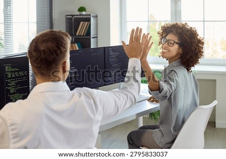 Developers teammates highfive after coding success. At dual monitors in a bright office, coders celebrate solved tasks and smooth teamwork. Focused work. Strong concept of teamwork and success.