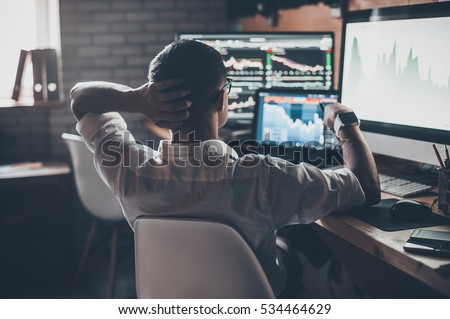 Developing new approaches. Rear view of young man in casual wear holding hand on the back of the head and working while sitting at the desk in creative office