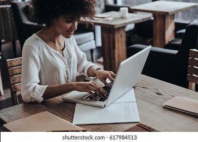 Developing New Project. Beautiful Young African Woman Using Computer And Smiling While Sitting In Cafe