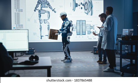 In Development Laboratory: Engineers Look At Robotics Exoskeleton Prototype Presentation With Person Testing It, Holding Heavy Carboard Box. Designing Wearable Exosuit To Help Warehouse Workers.