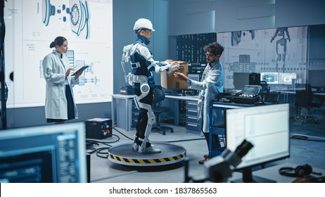 In Development Laboratory: Engineers Work On A Robotic Exoskeleton With A Person Testing It By Trying To Lift Heavy Cardboard Box. Designing Wearable Exosuit To Help Hard Labor Warehouse Workers