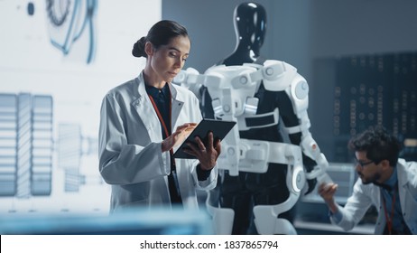 In Development Laboratory: Portrait Of A Beautiful Chief Female Engineer Using Digital Tablet Computer. Scientists Work On Prototype Exoskeleton Designed To Help Disabled People, Hard Labor Workers