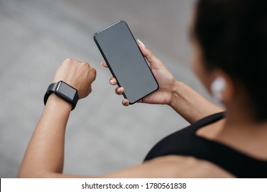 Device synchronization for sport. African american girl in sport bra and wireless headphones, looks at fitness tracker and smartphone in hands, free space