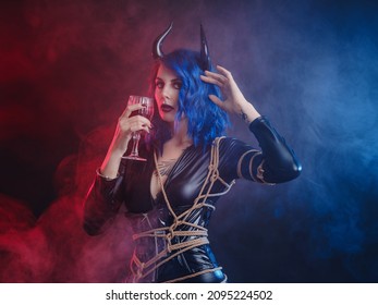Devil. Funny imp with black horns and latex suit with shibari at Halloween party. Halloween makeup. The devil costume. woman tastes red wine looks like blood in glass
