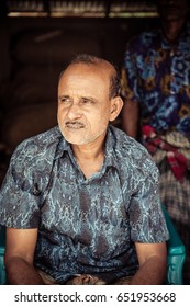 DHAKA, BANGLADESH - MAY 26, 2017: Portrait Of An Old Shop Keeper In Bangaldesh With A Bold Head And Wrinkled Face Sitting In A Plastic Chair With A Dark Background