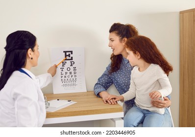 Diagnosis Of Child's Vision. Female Optometrist With Test Chart Checks Kid Eye Sight Who Came To Reception Together With Mom. Little Girl Sits In Her Mother's Arms And Calls Letters Shown By Doctor.