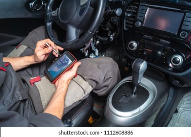 Diagnostics of car failures: an electrician in gray clothes is sitting in the car's interior and reading the trouble codes from a portable auto scanner with OBD2 interface.