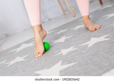 diagonal close-up two feet of a young woman, one foot standing on a green spiki massage ball on a gray carpet in a home interior.