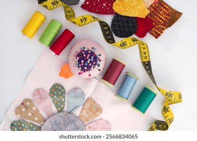 Diagonal flat lay ready to stitched flower applique,  twisted measuring tape, colorful spools of sewing and quilting thread, and straight Pins against white background 
