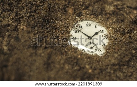 The dial of a mechanical watch sticks out of the ground. Found a clock in the sand on the beach. Bury time in the ground.