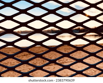 Diamond shaped diagonal forms are seen peering through a metal fence at the sunny river creating a geometric pattern background.