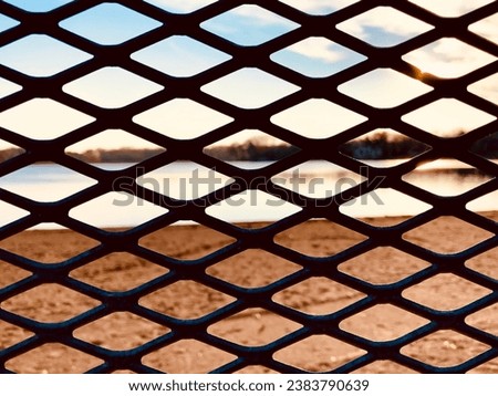 Diamond shaped diagonal forms are seen peering through a metal fence at the sunny river creating a geometric pattern background.