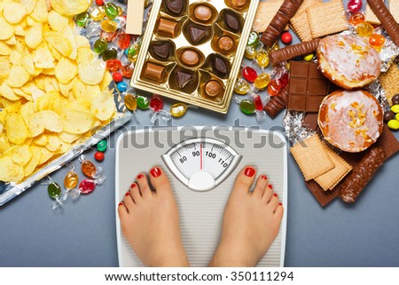 Diet concept. Feet of a young woman on bathroom scale and chocolate, jelly cubes, candies, chocolate bars, cookies, donuts, potato chips. Top view