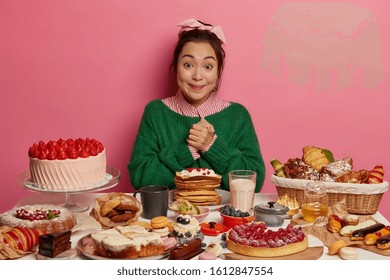 Diet Fail Concept. Cheerful Asian Woman Has Happy Reaction On Tasty Confectionery, Experiences Overeating, Clenches Hands Together, Has Cheat Meal Day, Sits At Big Table Against Pink Background