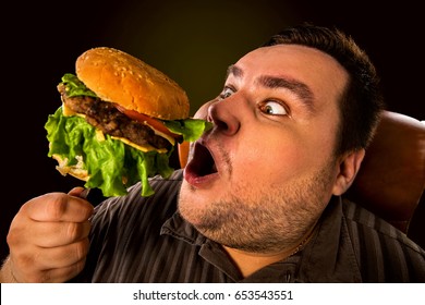 Diet failure of fat man eating fast food . Happy smile overweight person who crazy makes squint for fun eating huge hamburger on fork. Junk meal leads to obesity.