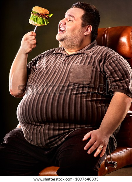 Diet failure of fat man eating fast food hamberger. Happy smile overweight person who In business chair eating huge hamburger on fork. Junk meal leads to obesity. Feast on occasion of feast.