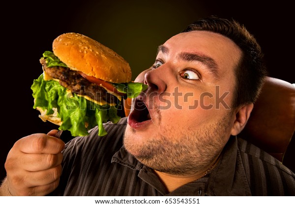 ファストフードを食べる太った男性の食事不良 フォークで巨大なハンバーガーを食べて楽しみに目をつぶる 狂った人は 太り過ぎの幸せなほほ笑みを浮かべる ジャンクミールは肥満につながる の写真素材 今すぐ編集