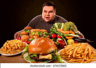 Diet fat man who makes choice between healthy and unhealthy food . Overweight male with hamburgers, french fries and vegetables trays trying to lose weight first time .Wide angle shooting.