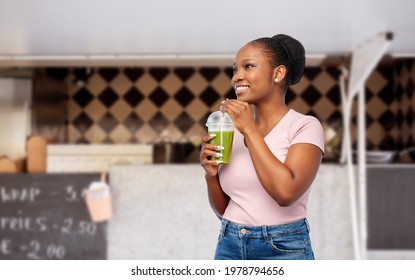 diet, healthy eating and detox concept - happy smiling young african american woman drinking green vegetable juice or smoothie from plastic cup with paper straw over food truck background - Powered by Shutterstock