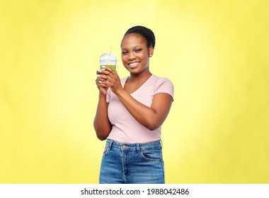 diet, healthy eating and detox concept - happy smiling young african american woman drinking green vegetable juice or smoothie from plastic cup with paper straw over illuminating yellow background - Powered by Shutterstock