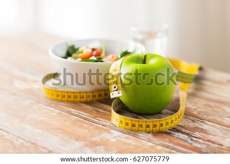 diet, healthy eating, food and weigh loss concept - close up of green apple and measuring tape with salad on wooden table