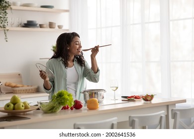 Diet, Healthy Lifestyle. Cook At Home, Household And Nutrition. Glad Young African American Woman Cooks Soup And Tastes Food In Modern Kitchen Interior With Bright Vegetables, On Window Background