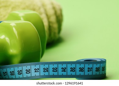 Diet And Sport Regime Concept. Sports Regime Idea, Defocused. Dumbbells In Bright Green Color, Twisted Measure Tape And Towel On Green Background. Tape Measure In Cyan Blue Color By Barbells, Close Up